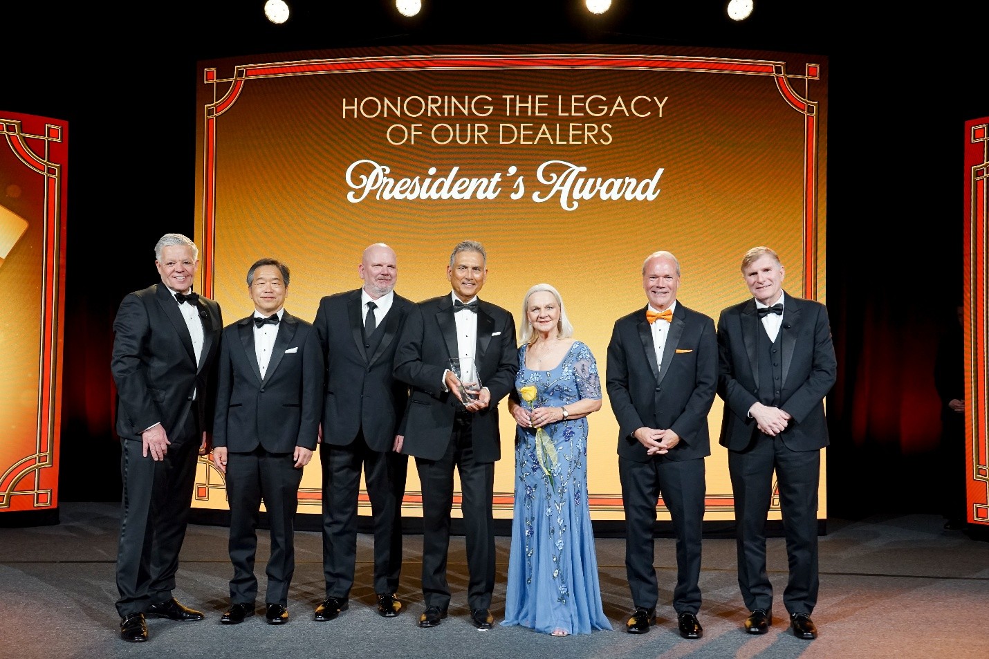 Seven people in formal evening wear on a stage, posing with a trophy and a yellow rose under a backdrop that reads 'President's Award' and 'Honoring the Legacy of Our Dealers'