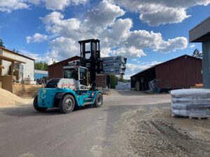 Blue forklift in an outdoor industrial yard carrying wrapped pallets, with warehouses and stacked materials nearby under a cloudy sky